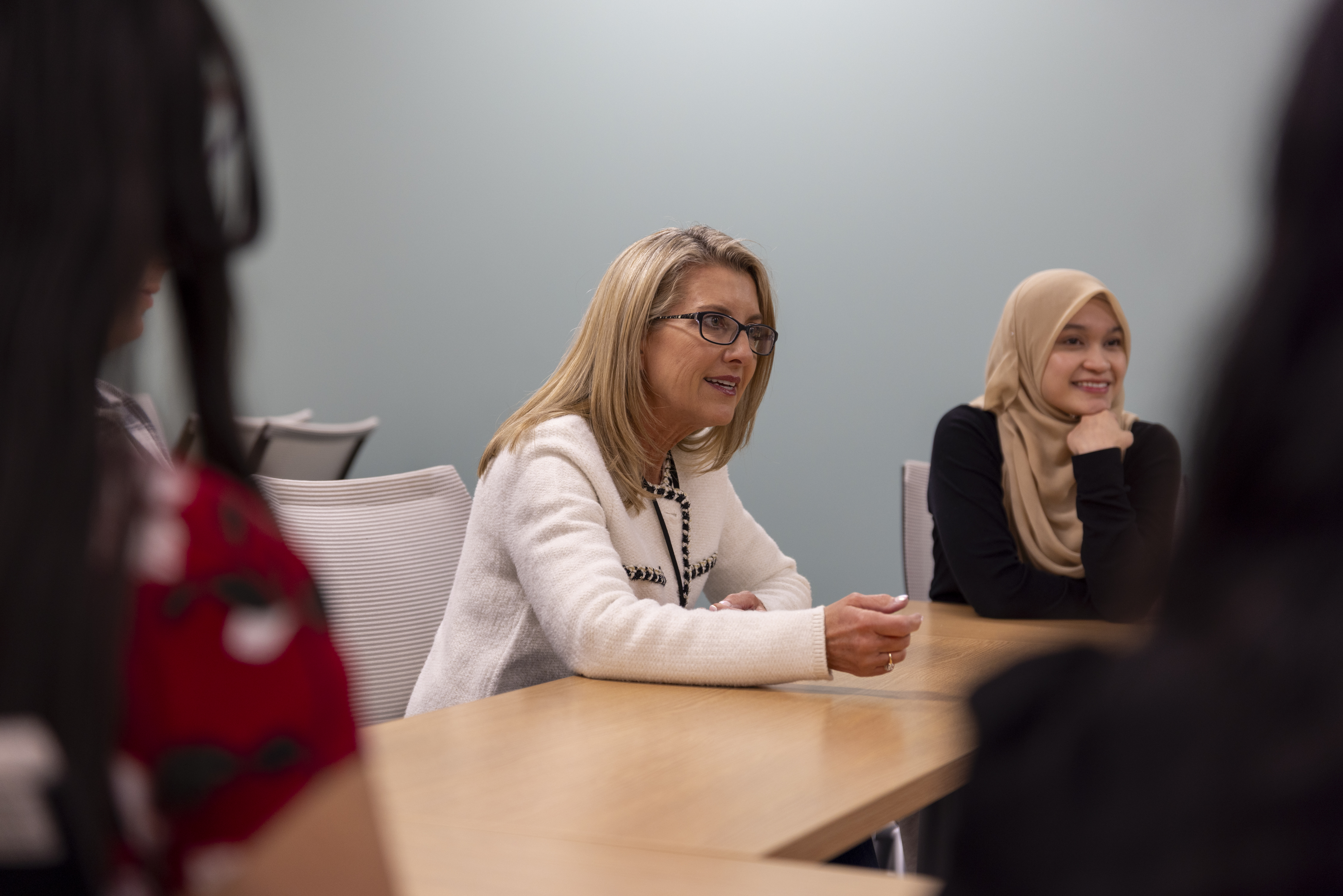 A white woman sits at a table leading a group in discussion. She sits next to a woman wearing a hijab.