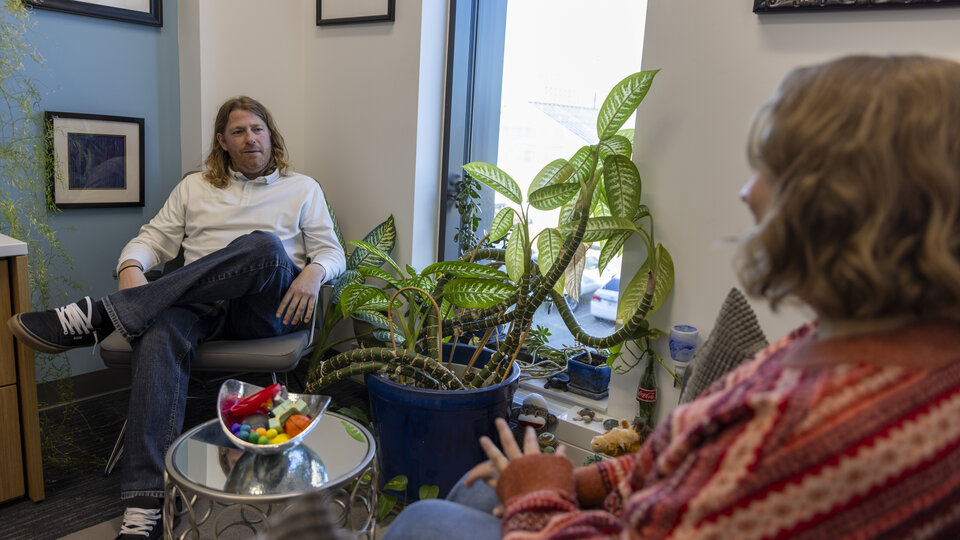 A white male counselor in an office with plants and fidget toys sits in a chair across from a white female student, conducting a one-on-one counseling session.