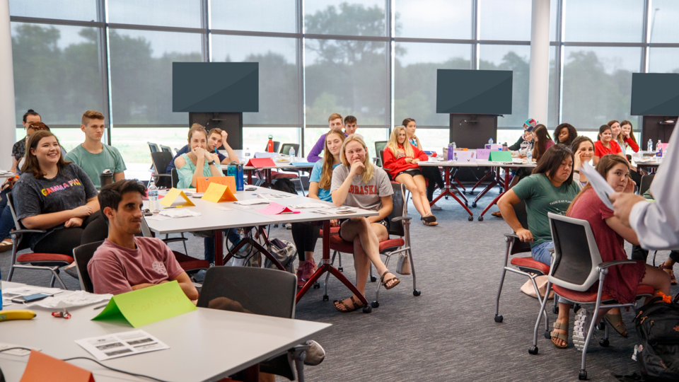 Students grouped at tables listen to a speaker