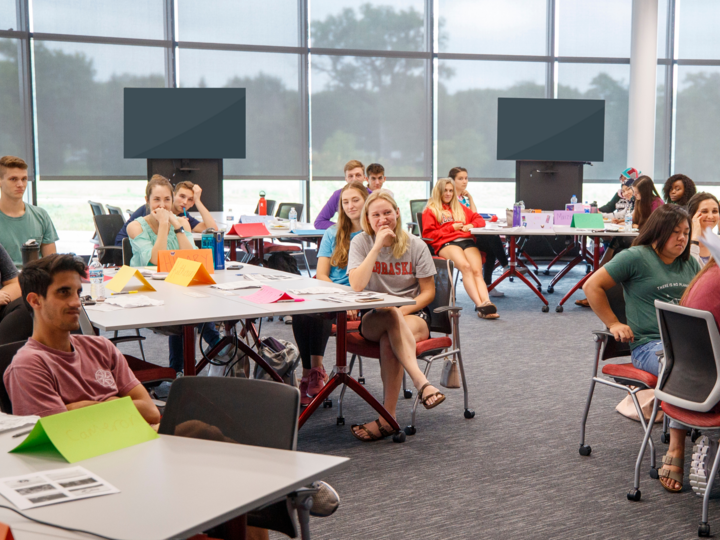 Students grouped at tables listen to a speaker