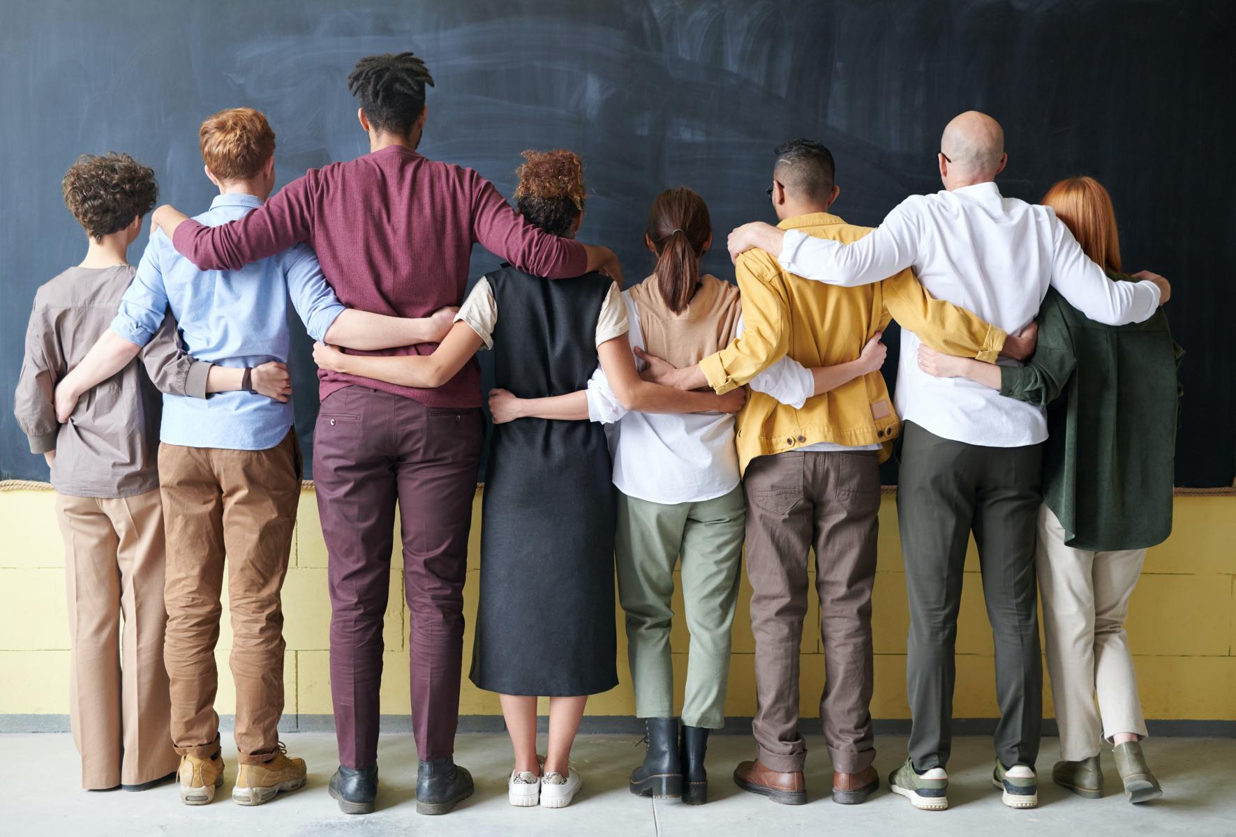 A group of diverse individuals with their backs to the camera and their arms around one another facing a chalkboard